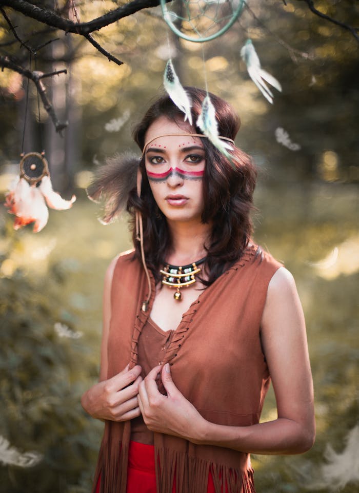 A woman in tribal-inspired attire and face paint poses outdoors surrounded by dreamcatchers.